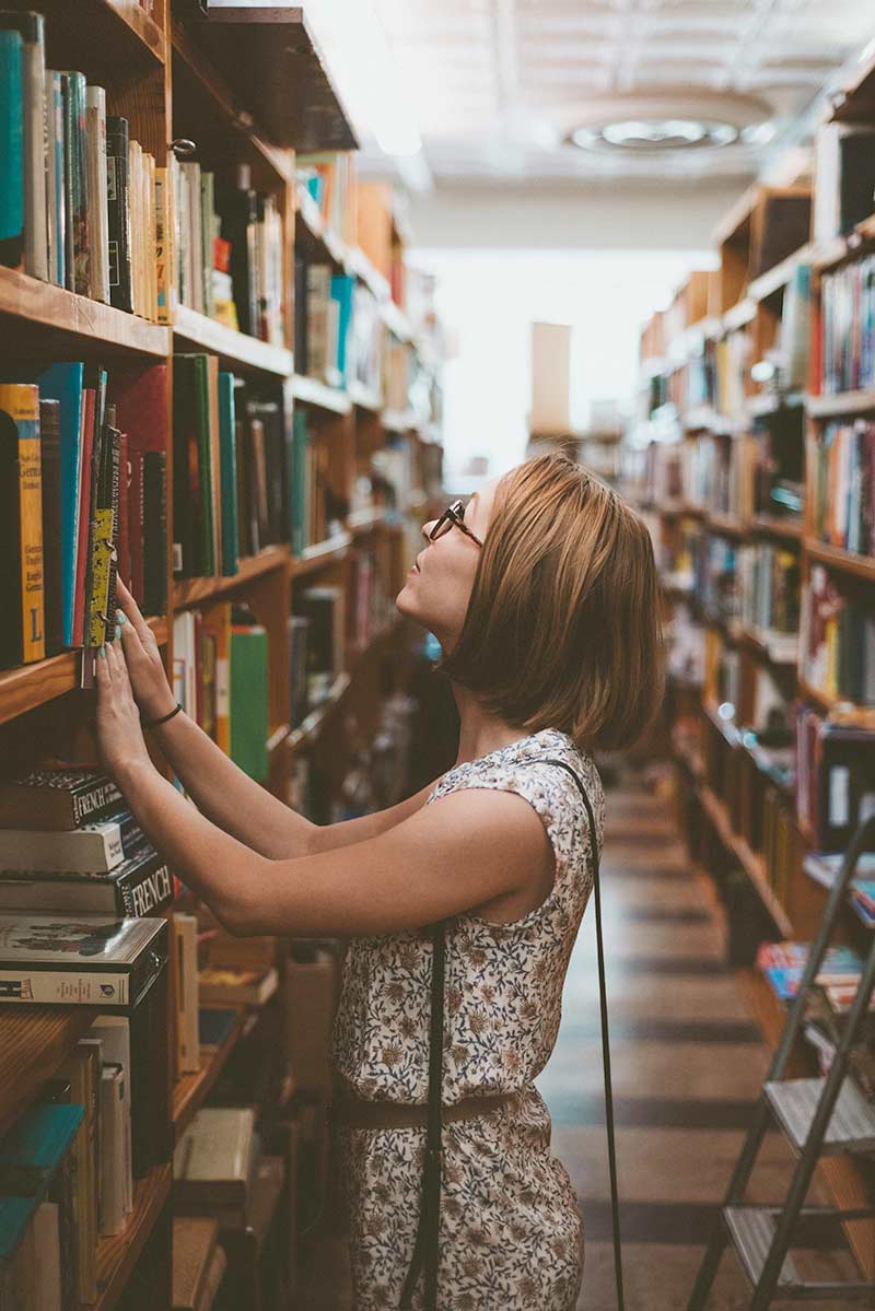 A woman in a library, intently searching for books on the shelves filled with various literary works