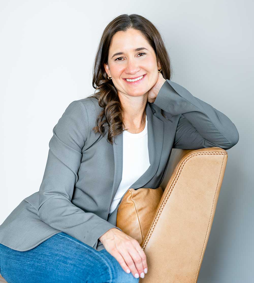 Smiling woman in grey blazer and jeans, seated in a brown chair.