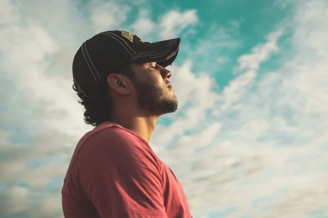 Man standing outdoors with eyes closed, taking a deep breath under a bright sky, representing stress relief and mental calm.