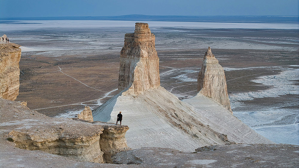 A person standing on a rocky desert cliff overlooking a vast, rugged landscape.